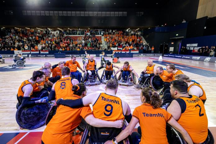 Wheelchair rugby players wearing orange jersey are forming a circle on the court before a game